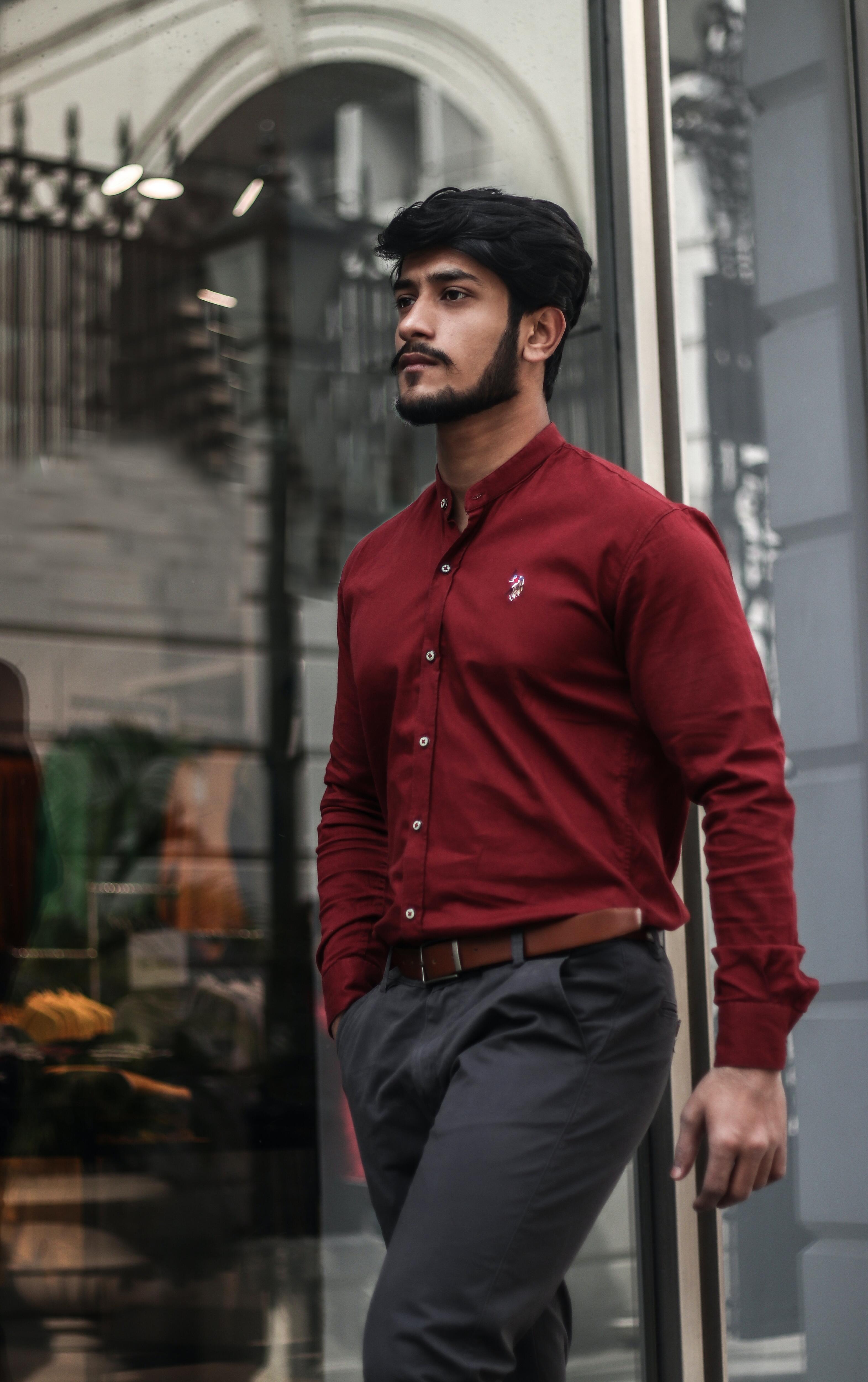 Man with neatly-trimmed facial hair in red button-up shirt and dark pants with belt walks along a street