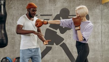 A man in a skull cap instructs a woman in boxing gloves on punching technique