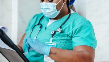 Black nurse in scrubs and mask with stethoscope making notes on a chart